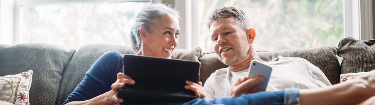A couple in their 50's relax in their home on the living room couch, enjoying reading and surfing the internet on their mobile touchscreen phones and computer tablet