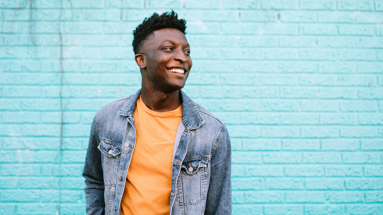 A smiling African American man stands in front of a blue wall wearing nice casual clothing. Positive lifestyle portrait. Shot in Portland, OR