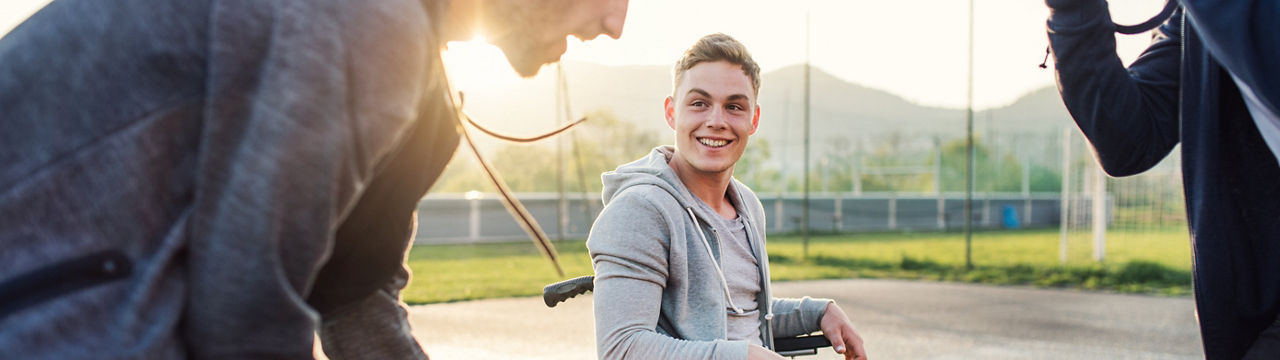 A boy in wheelchair with two teenager friends playing basketball outside at sunset. Close up.