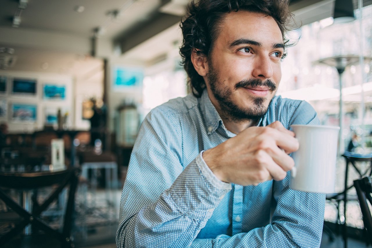 Young man sitting in cafeteria holding a cup of coffee and looking away, with copy space