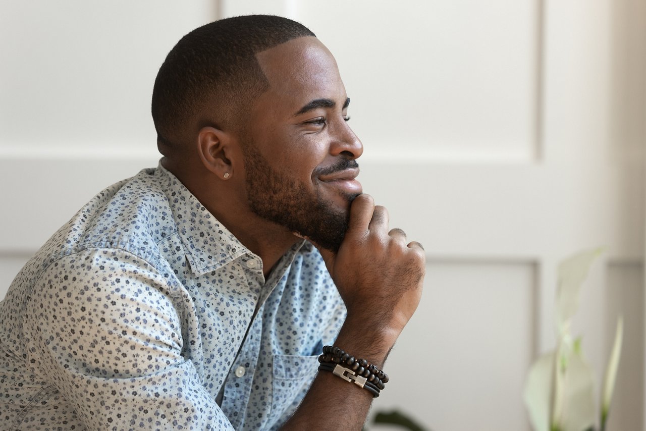 Dreamy calm African American young man looking away contemplating daydreaming sitting at home
